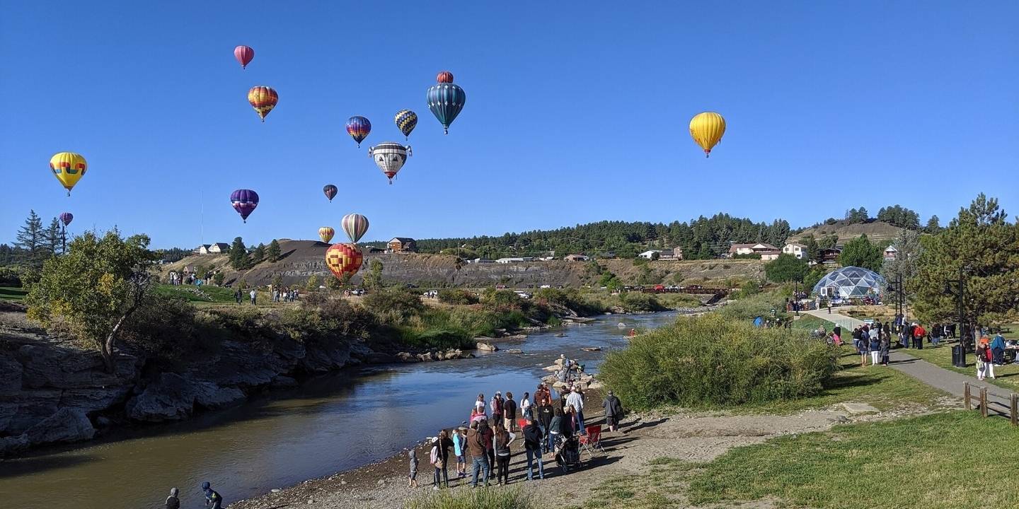 A crowd of people along the San Juan River looking up at a group of hot air balloons in the sky during Pagosa Colorfest