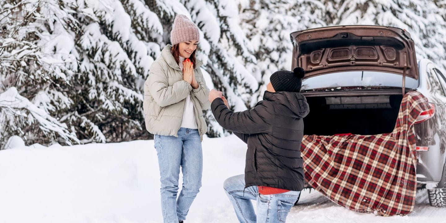 A man kneels in the snow, proposing to a woman with a ring, surrounded by a winter forest